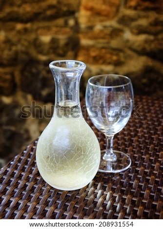 Ornate pitcher with cold white wine and one empty glass on wicker table in front of rough stone wall in rustic restaurant. Reflection. A game of light and shadow. Selective focus on the pitcher.