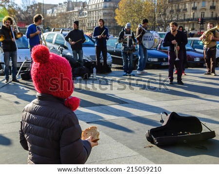 PARIS, FRANCE - DECEMBER 8, 2013: Brass band performs and unidentified little girl  stands close to a cover with coins, holding slice of bread, creating by a chance \