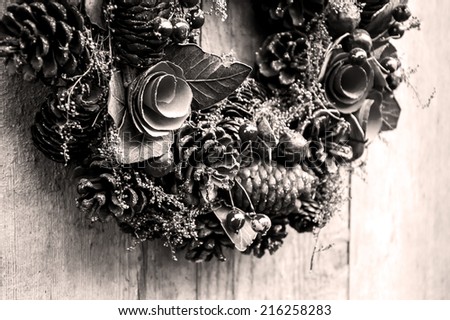 Vintage Christmas wreath with pine cones, flowers and red berries hanging on the grungy wooden door.  Greeting card. Aged photo. Black and white.