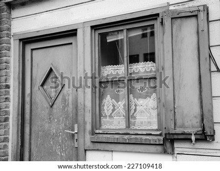 A front of house. A door and a window with lace curtain decorated with windmill and tree pattern and wooden hinged shutters. (Amiens, Picardy, France)  Rustic background. Aged photo. Black and white.