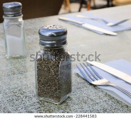 Salt cellar, pepper caster, forks and knives on granite table at an outdoor restaurant.