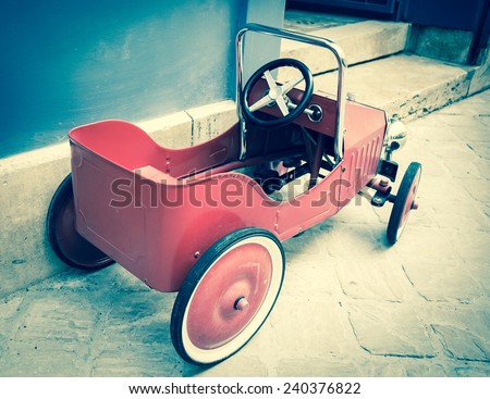 Red vintage toy car near entrance to the house. Back and side view. Aged photo.