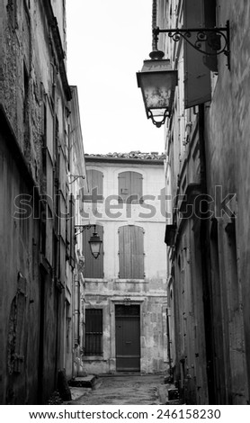 Narrow the street in Arles (Provence, France). Weathered stucco walls, red wooden shutters and forging lanterns. Aged photo. Black and white.