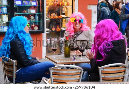 VENICE, ITALY - FEBRUARY 14, 2015: Three young women in funny glasses (in shape of word COOL) and colorful wig drinking red wine in cafe during Carnival. Traditional Carnival in Venice is annual event