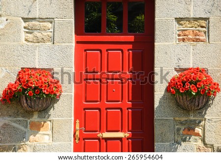 Stone house wall with red wooden door (entry to the manor\'s garden) and and hanging pots with red geranium flowers. Brittany, France.
