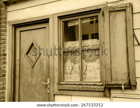 A front of the house. A door and a window with white lace curtain decorated with windmill and tree pattern and wooden hinged shutters . (Amiens, Picardy, France)  Rustic background. Aged photo. Sepia.