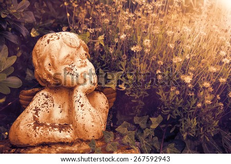 Weathered statue of an infant angel in overgrown garden. Sunset golden light. Toned photo.