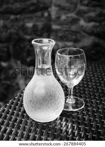 Ornate pitcher with wine and one empty glass on wicker table in front of rough stone wall in rustic restaurant. A game of light and shadow. Selective focus on the pitcher. Aged photo. Black and white.