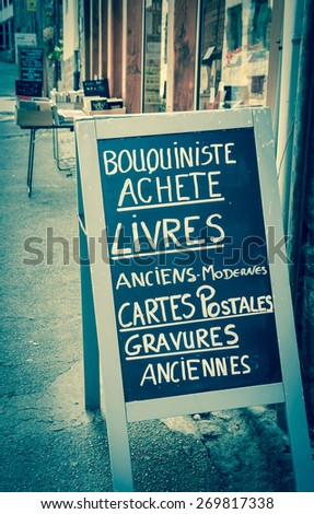 Signboard outside old books store in France. Text in French meaning: Secondhand bookseller buying old and modern books, postcards, ancient prints. Aged photo.