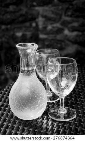 Ornate pitcher with wine and two empty glasses on wicker table against rough stone wall in rustic restaurant. A game of light and shadow. Aged photo. Black and white.