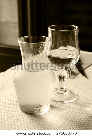 Misted pitcher with cold white wine and one glass on paper tablecloth in simple rustic restaurant. Aged photo. Sepia.
