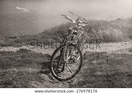 Two bicycles on  hill over the sea in sunny day and a boat sailing at background. Breton coast near Cap de la Chevre. Brittany, France. Sunlight rays. Aged photo. Black and white.