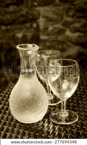 Ornate pitcher with wine and two empty glasses on wicker table against rough stone wall in rustic restaurant. A game of light and shadow. Aged photo. Sepia.