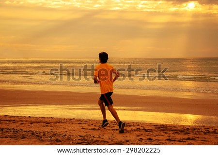 Young man running on beach at sunset. Back view. Dramatic sky lighting with sun rays glowing through the clouds. A game of light and shadow. Toned photo.