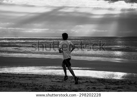 Young man running on beach at sunset. Back view. Dramatic sky lighting with sun rays glowing through the clouds. A game of light and shadow. Aged photo. Black and white.
