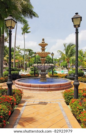 Tropical gardens with a beautiful fountain and ponds flanked by ornate hedges and lamposts.