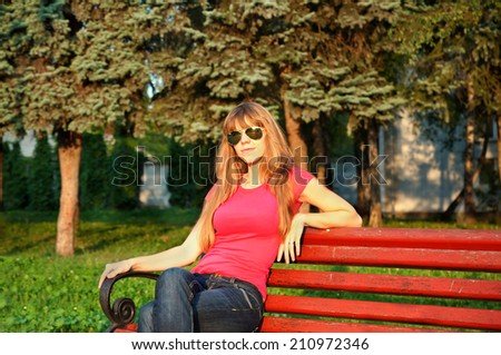 beautiful young woman in sunglasses and jeans sitting on the bench at the park and enjoying sunset