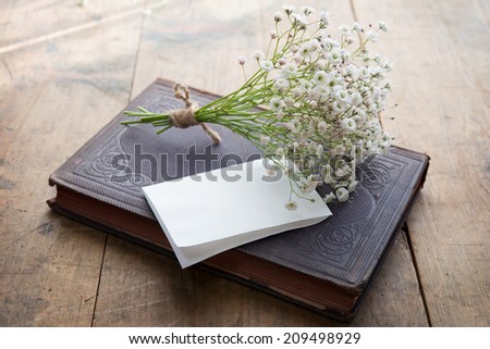 Vintage book and a small bouquet of baby's breath flowers and a blank sheet of folded white paper, on a well used old desk or wooden surface.