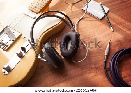 Guitar recording scene. An electric guitar, memo pad , and a professional grade headphones on a rustic or bare wooden table, with by-the-window type warm light coming in.