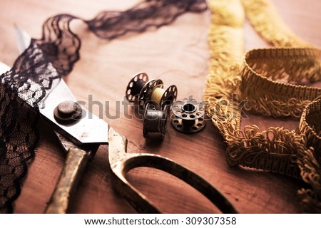 Fringe or lace tapes and silk trimmings with gold (brass) scissors and old sewing machine bobbins on a old grungy work table. Tailor\'s workbench. textile or fine cloth making.Shallow depth of field.