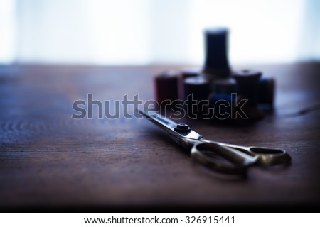 Well used tailor\'s scissors, with sewing  thread wooden reels or bobbins in background, on a old grungy work table. Shallow depth of field. Intentionally shot  in low key and nostalgic muted tone.