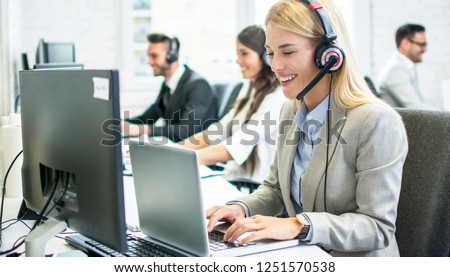 Beautiful blonde customer support female telephone worker using laptop and talking with client at call center