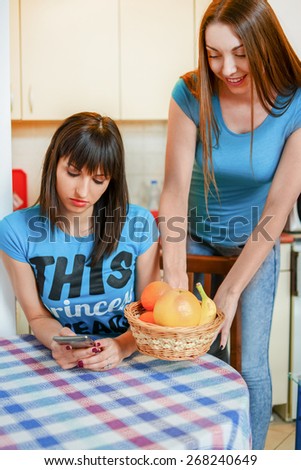 Young woman bringing fruits while her female friend using smartphone at home.