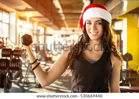 Young woman in Santa hat holding dumbbell at gym. New Year and Xmas concept.