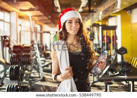 Young woman with red Santa hat, towel and water bottle in gym. Snow effect on photo.