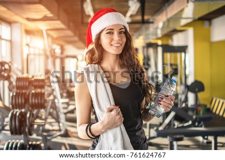 Young fir woman with red Santa hat, towel and water bottle in gym. New Year. Christmas, holidays, fitness, and gym concept.