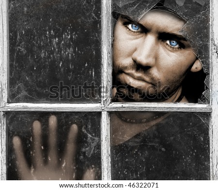 Handsome young Man with piercing Blue Eyes stares through dirty old broken Window with his Hand pressed against the Glass with room for a message above