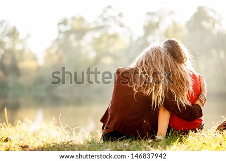 Two Young Women Sitting On Grass Hugging Rear View