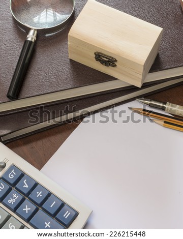 Pen and pencils on blank paper with magnifying glass and wooden box on stack of books.