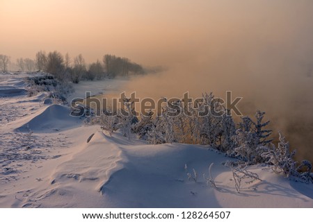 Fog over the river. On the bank of the river grass and trees covered with snow. Fog lights illuminated the setting sun.