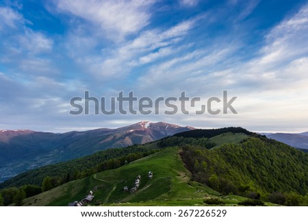 Landscape with mountain ranges, forested slopes, mountain pasture with buildings summer farm, snowfields on top of the ridge and sky during sunrise. Carpathians.