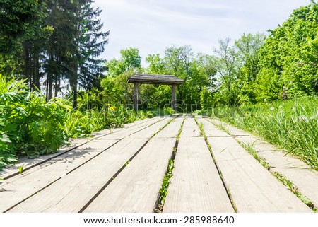 Garden path, lined with wooden planks in the grass, leading to a decorative wooden gate with a roof.