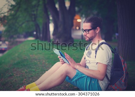 The young man, bearded student sitting in a park on the nature of reading or working on the tablet.