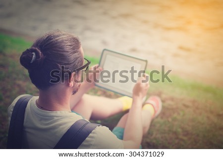 The young man, bearded student sitting in a park on the nature of reading or working on the tablet.