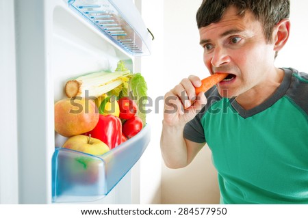 Man eating carrot near the opened refrigerator