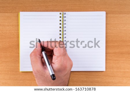 Hand  holding black pen on white, open note paper on wood background.