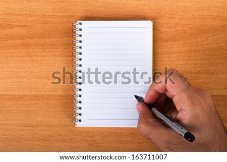 Hand holding black pen for writing to blank, white notebook on wood background.
