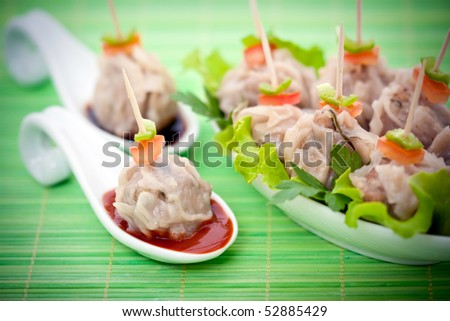 Chinese steamed meat dumplings with sauce on ceramic spoon and on a garnished plate sitting on a green bamboo mat