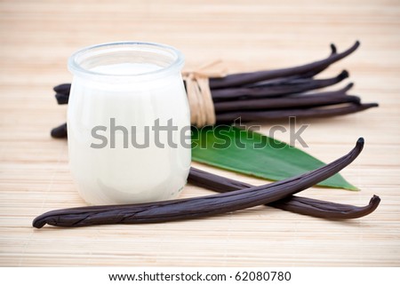 Glass container of yogurt with fresh vanilla beans on leaf and bamboo mat.