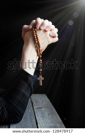 Hands holding rosary with a cross and folded in prayer in the rays of light on the wooden table