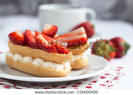 Fresh cream eclairs with whipped cream and strawberries on white plate and cup of coffee on white wooden table, selective focus