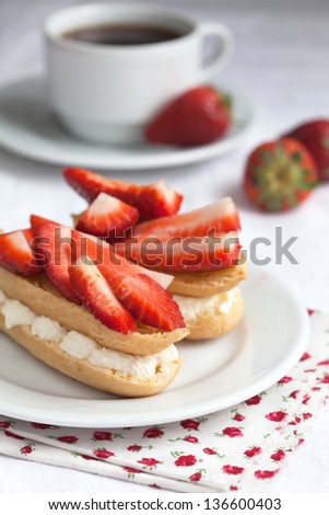 Fresh cream eclairs with whipped cream and strawberries on white plate and cup of coffee on white wooden table