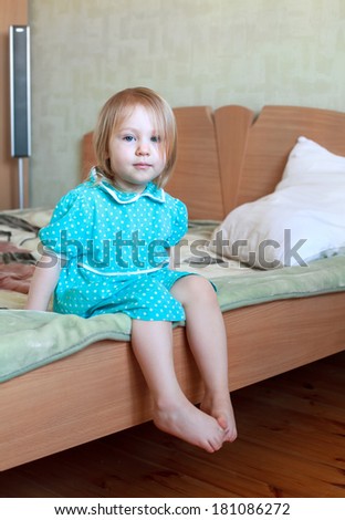 Small sad blonde girl in blue dress sitting on the bed dangling her legs, selective focus on face