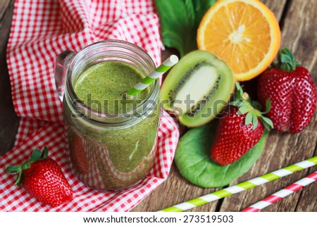 Healthy green smoothie made from spinach, kiwi, strawberries and oranges in a jar with green straw on a wooden table, selective focus