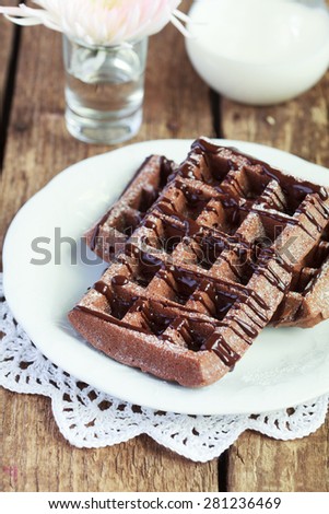 Homemade chocolate waffles with cocoa  from whole wheat flour, decorated with melted chocolate and powdered sugar on a white plate and a pitcher of milk on a wooden table, selective focus