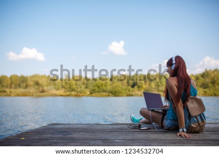 Photo on back of young brunette in headphones with backpack and laptop in hands sitting on river bank